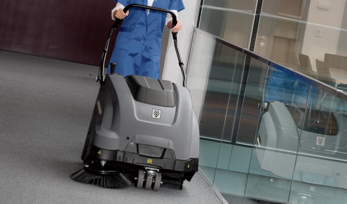Profesionální ruční zametací stroj KM 75/40 W Bp od Karcher s elektrickým motorem. Profesionální ruční zametací stroj KM 75/40 W Bp od Karcher s elektrickým motorem. Žena zametá chodbu v kancelářské budově pomocí profesionálního zametacího stroje KM 75/40 W Bp vyráběného firmou Karcher.