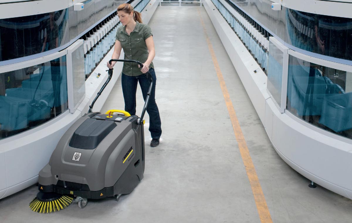 Profesionální ruční zametací stroj KM 75/40 W Bp od Karcher s elektrickým motorem. Profesionální ruční zametací stroj KM 75/40 W Bp od Karcher s elektrickým motorem. Žena zametá výrobní halu pomocí profesionálního zametacího stroje KM 75/40 W Bp vyráběného firmou Karcher.